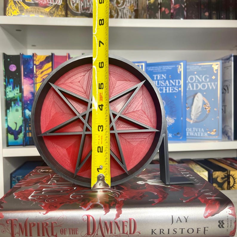 May include: A red and black bookend with a 7-pointed star design. The bookend is 5 inches tall and is sitting on top of a book titled "Empire of the Damned" by Jay Kristoff.