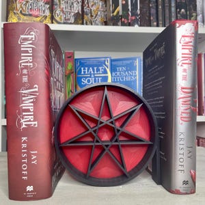 May include: A red and black 8-pointed star bookend. The bookend is in front of two copies of the book "Empire of the Damned" by Jay Kristoff.