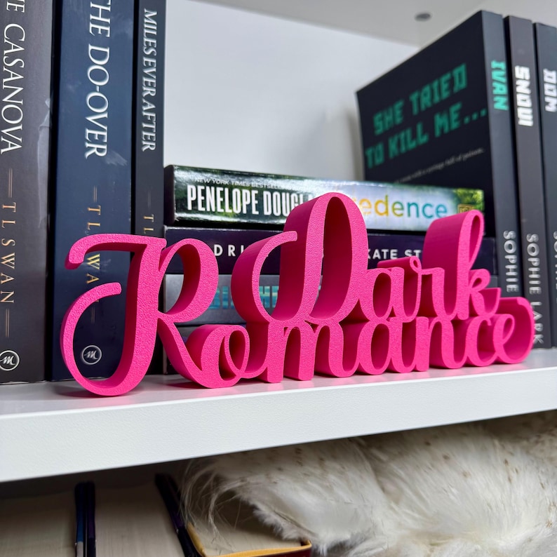 Puede incluir: Un letrero de madera rosa con las palabras "Dark Romance" en una fuente cursiva. El letrero est&aacute; sentado en una estanter&iacute;a blanca con otros libros detr&aacute;s.