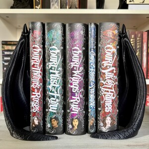May include: A set of five fantasy novels with black covers and silver lettering. The titles are "A Court of Thorns and Roses", "A Court of Mist and Fury", "A Court of Wings and Ruin", "A Court of Frost and Starlight", and "A Court of Silver Flames". The books are displayed on a bookshelf with a black dragon bookend.