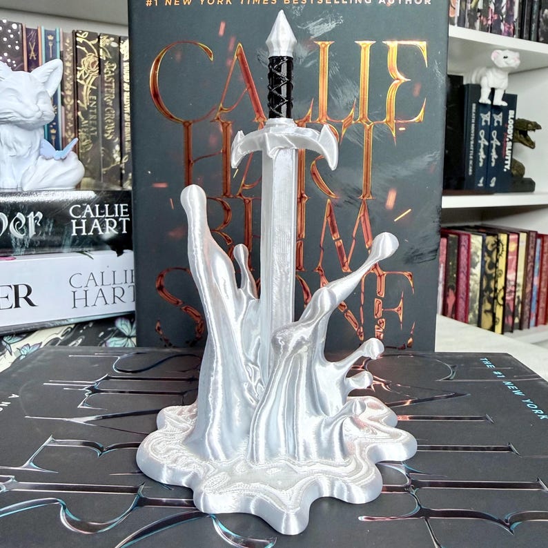 May include: A silver-colored sword bookend with a black handle, emerging from a stylized splash. The bookend is in front of a book with the title "CALLIE HART" and the text "#1 NEW YORK TIMES BESTSELLING AUTHOR".