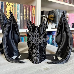 May include: Black 3D printed dragon head bookends with detailed scales and wings. The dragon head is in the center with two wings on either side.