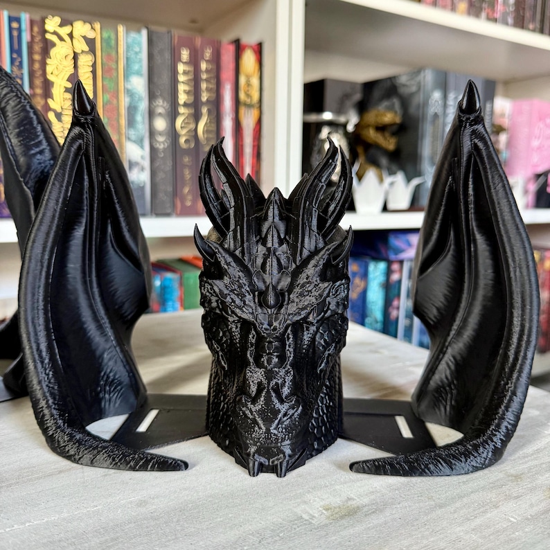 May include: Black 3D printed dragon head bookends with detailed scales and wings. The dragon head is in the center with two wings on either side.