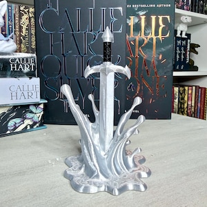 May include: A silver-colored sword sculpture with a black handle, set in a stylized water splash base. The sculpture is in front of books with the author's name, Callie Hart, and book titles visible.