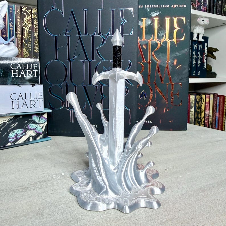 May include: A silver-colored sword sculpture with a black handle, set in a stylized water splash base. The sculpture is in front of books with the author's name, Callie Hart, and book titles visible.
