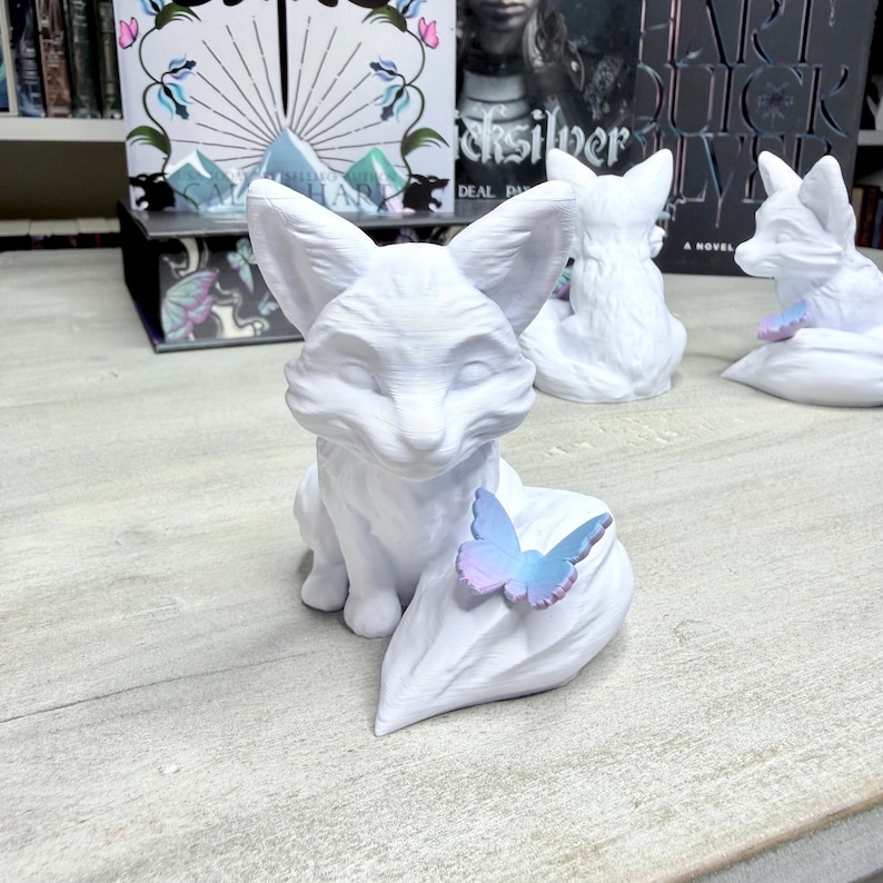 May include: A collection of white fox figurines, each with a stylized design. One fox has a butterfly with blue and purple hues resting on its tail. The figurines are displayed on a light-colored surface, with books and decorative items in the background.