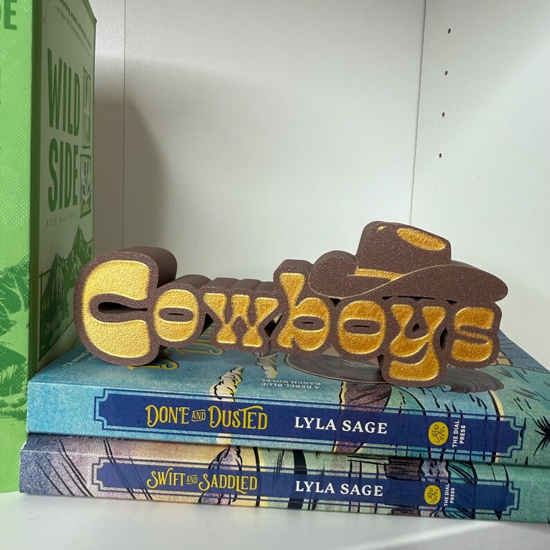 Puede incluir: Un letrero decorativo marr&oacute;n y dorado que dice "Cowboys" con un sombrero de vaquero. El letrero est&aacute; colocado encima de dos libros titulados "Don't and Dusted" y "Swift and Saddled" de Lyla Sage. Un libro verde es visible a la izquierda.
