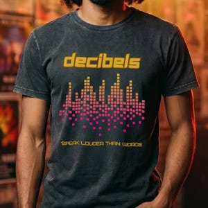 May include: Dark gray t-shirt with the word "decibels" in gold lettering above a pink and gold graphic of sound waves. The phrase "Speak Louder Than Words" is printed below the graphic. The t-shirt is worn by a person with dark hair.