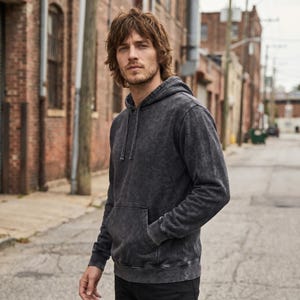 May include: A dark gray hoodie with a kangaroo pocket and drawstring hood. The hoodie has a faded, textured appearance. The person is wearing black pants and standing on a paved road in front of brick buildings.
