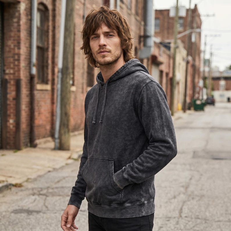 May include: A dark gray hoodie with a kangaroo pocket and drawstring hood. The hoodie has a faded, textured appearance. The person is wearing black pants and standing on a paved road in front of brick buildings.