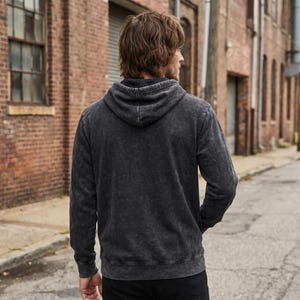 May include: A dark gray hooded sweatshirt with a faded, textured appearance. The hoodie has a drawstring and a kangaroo pocket. The person is wearing black pants. The background is a brick building and a paved street.