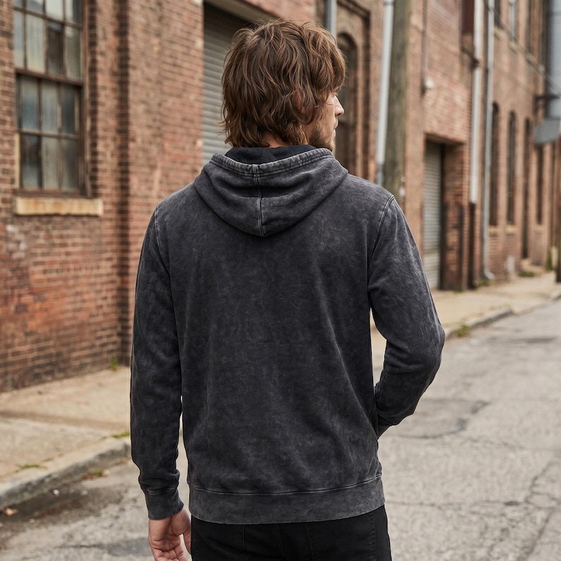 May include: A dark gray hooded sweatshirt with a faded, textured appearance. The hoodie has a drawstring and a kangaroo pocket. The person is wearing black pants. The background is a brick building and a paved street.