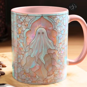May include: A ceramic mug with a pink handle and interior. The exterior features a stained-glass design with a ghost figure in the center. The color palette includes pastel shades of pink, blue, and peach. The mug is on a wooden surface.