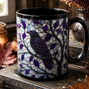 May include: A black ceramic mug featuring a stained glass style illustration of a dark raven perched on a tree branch with purple leaves. The mug has a glossy black handle and rim, showcasing a detailed mosaic pattern of light and dark tones.