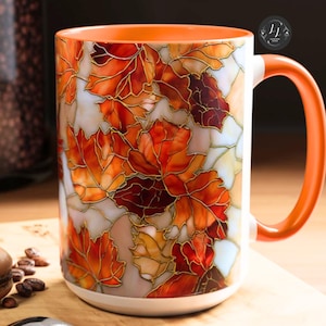 May include: A white ceramic mug with an orange interior and handle, decorated with a stained-glass style design of autumn leaves in shades of red, orange, and brown. A spoon and a macaron are on a wooden surface.