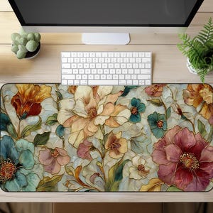 May include: A floral-patterned desk mat featuring a colorful design of large flowers in shades of orange, pink, blue, and cream. The mat has a black border and is placed on a light wood surface, with a keyboard and monitor visible.