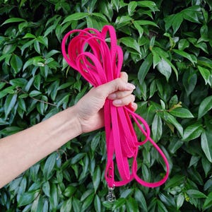 Puede incluir: Una correa rosa brillante para perros con un cierre plateado y un asa a juego. La correa se sostiene en una mano sobre un fondo de hojas verdes. La correa está hecha de un material suave y flexible.