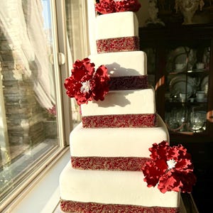 May include: A white square wedding cake with red sugar flowers and gold and red ribbon accents.
