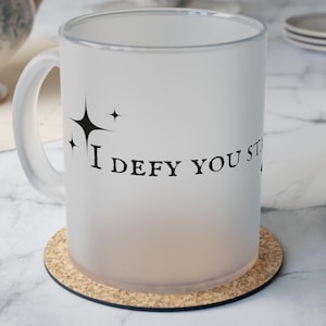 May include: A white frosted mug with a cork coaster. The mug has a black design that says "I defy you still" with three stars above the text.