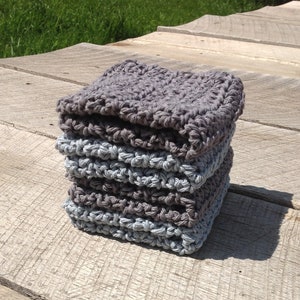 May include: A stack of four square, hand-crocheted washcloths in shades of gray. The washcloths are stacked on a weathered wooden surface, with the top cloth a darker gray than the others. The texture of the crochet is visible.