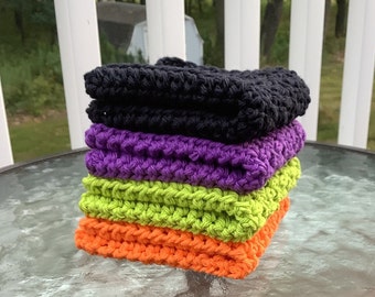 May include: A stack of five crocheted washcloths in Halloween colors: black, purple, green, and orange. The washcloths are stacked on top of each other, with the black cloth on top and the orange cloth on the bottom. The washcloths are made of a thick, textured yarn.