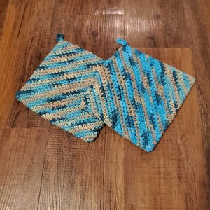 May include: Two handmade, square crochet potholders. The potholders feature a striped pattern in shades of blue, teal, and tan. Each potholder has a small loop for hanging. The potholders are displayed on a wooden surface.