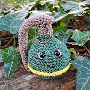 May include: A green crocheted grenade with a brown crocheted handle and a yellow crocheted band. The grenade has a smiling face with black eyes and a black stitched mouth.