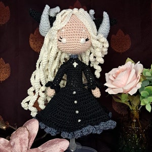 Könnte beinhalten: Eine gehäkelte Puppe mit weißen Haaren, schwarzen Hörnern und einem schwarzen Kleid mit weißen Knöpfen. Die Puppe steht auf einem durchsichtigen Plastikständer. Die Puppe befindet sich vor einem braunen Hintergrund mit rosa Blumen.