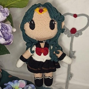 Könnte beinhalten: Eine gehäkelte Puppe mit grünen Haaren, einer schwarz-weißen Matrosenuniform und einer roten Schleife. Die Puppe hält einen silbernen herzförmigen Zauberstab mit einem roten Edelstein.