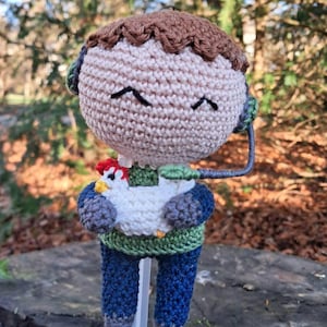 Puede incluir: Un juguete de amigurumi de crochet que representa a una persona con una camisa verde y pantalones azules, sosteniendo un pollo blanco de crochet. El juguete tiene el pelo marrón y un cordón negro sujeto a su cabeza.