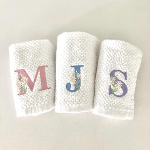 May include: Three white, rolled washcloths with embroidered floral initials. The letters are pink, blue, and purple, each with a floral design. The washcloths have a textured, waffle-weave pattern.