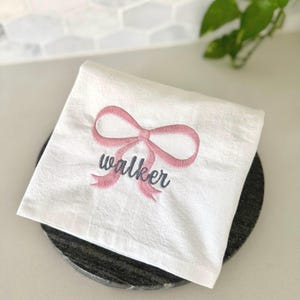 May include: White hand towel with a pink embroidered bow and the name "walker" in gray script. The towel is folded and placed on a dark gray, round coaster. The background is a light-colored countertop and a white tile backsplash.