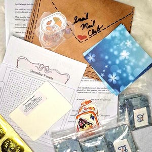 May include: A selection of stationery items, including a brown envelope with "Snail Mail Club" written on it, stickers, and small cross-stitch pieces. There are also journaling prompts and a blue floral card. The items are arranged on a white surface.