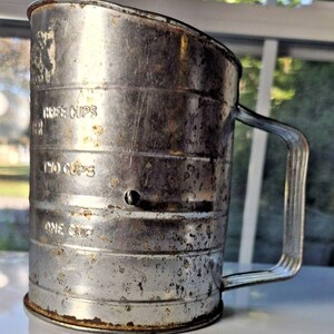 May include: Vintage metal flour sifter with a handle. The sifter has measurements for one, two, and three cups. The metal has a worn, rustic appearance with signs of age and use. The handle is a simple, curved design.
