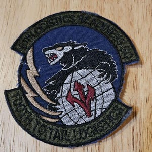 Op de afbeelding: Geborduurde patch met een wolf over een wereldbol, met rode pijlen. De patch heeft tekst rond de rand en een bliksemschicht. De patch ligt op een houten oppervlak.