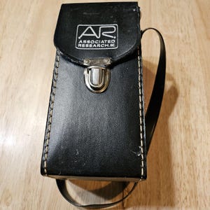 May include: Black leather case with a flap closure and a shoulder strap. The case features the text "AR ASSOCIATED RESEARCH, INC." in white. The case is stitched with white thread and has a metal clasp.
