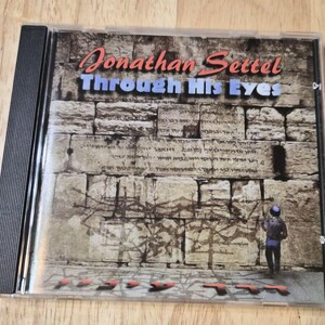 May include: A CD case featuring the album "Through His Eyes" by Jonathan Settel. The cover art depicts a stone wall with a person standing in front of it. The artist's name is in red and the album title is in blue.