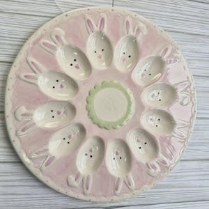 May include: A pink ceramic Easter egg serving platter with bunny-shaped egg holders. The platter has a scalloped edge and a central circular area. The bunny faces have black dot eyes and pink accents.
