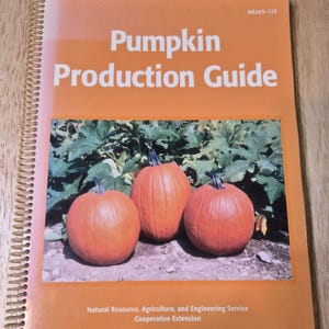May include: A spiral-bound book titled "Pumpkin Production Guide" with an orange cover. The cover features a photo of three orange pumpkins in front of green foliage. The book is a guide from the Natural Resource, Agriculture, and Engineering Service.