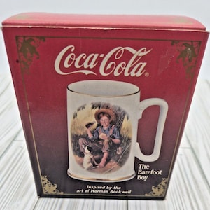 May include: A Coca-Cola mug featuring an illustration of a barefoot boy with a dog, inspired by Norman Rockwell's art. The mug has a white background with a gold rim and a white handle. The mug is in a red box with the Coca-Cola logo.