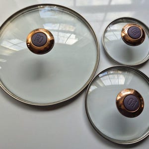 May include: Three clear glass pot lids with silver rims and copper-colored handles. The handles have a purple center with the letters "PD" embossed. The lids are different sizes, suitable for various pots and pans.