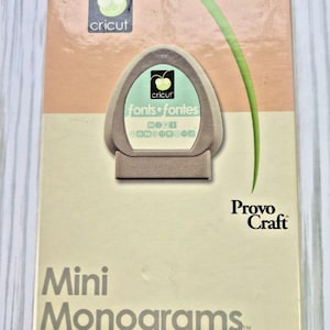May include: A Cricut Mini Monograms font cartridge box. The box is peach and cream-colored with the Cricut logo and the words "fonts fontes" and "Mini Monograms". The box also has the Provo Craft logo.