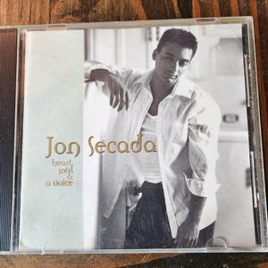 May include: A CD case featuring Jon Secada. The cover is in black and white, with Secada leaning against a wall. The album title "heart, soul & a voice" is printed in gold lettering.