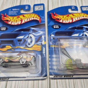 May include: Two Hot Wheels toy packages featuring blue backing cards with flame logos. The left package contains a yellow and black Deora model car. The right package contains a silver-colored scooter toy encased in a clear plastic blister pack.