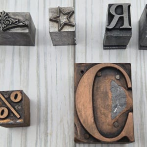 May include: A collection of six metal letterpress printing blocks. The set includes a rectangular block with a decorative filigree design, a square block with a star, two blocks with the letters R and P, a small square block with a percent sign, and a large rectangular block featuring a prominent copper-colored letter C.