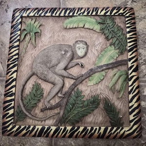 May include: A square, relief-style wall art piece featuring a monkey perched on a tree branch amidst lush green foliage. The monkey is gray, and the background is a light brown. The frame has a zebra print pattern.