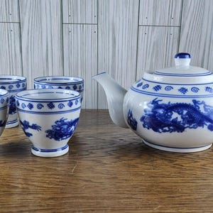May include: A white porcelain tea set with blue dragon designs. The set includes a teapot with a lid and four matching teacups. The tea set is displayed on a wooden surface.