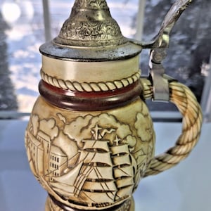 May include: A vintage ceramic beer stein with a detailed nautical scene featuring sailing ships and buildings. The stein has a metal lid and handle, with a rope-like design on the handle. The base reads "Bark T. Stausbol".
