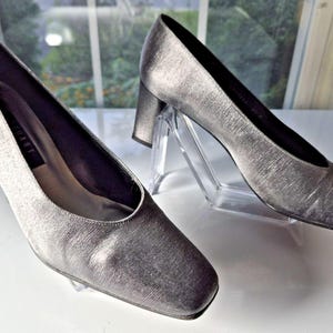 May include: A pair of silver-colored, heeled pumps. The shoes have a square toe and a block heel. The pumps are displayed on clear plastic stands. The shoes appear to be made of a textured fabric.