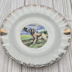 May include: A round white porcelain ashtray featuring a central illustration of a brown and white hunting dog in a field with a bird in flight. The rim is scalloped with a metallic gold trim and two integrated notches for resting cigarettes.
